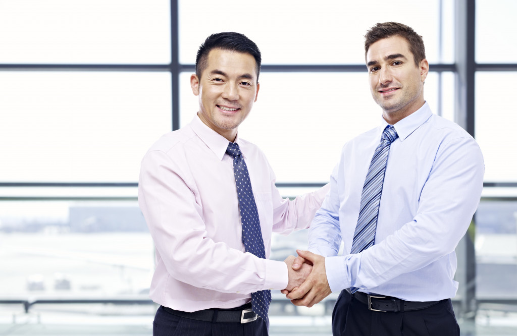 two businessmen, one asian and one caucasian, shaking hands looking at camera at modern airport.