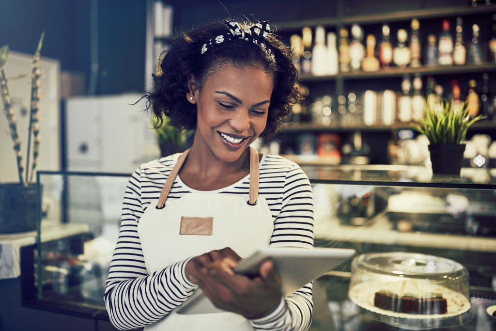 African entrepreneur wearing an apron and working with a digital tablet while standing in front of the counter of her trendy cafe