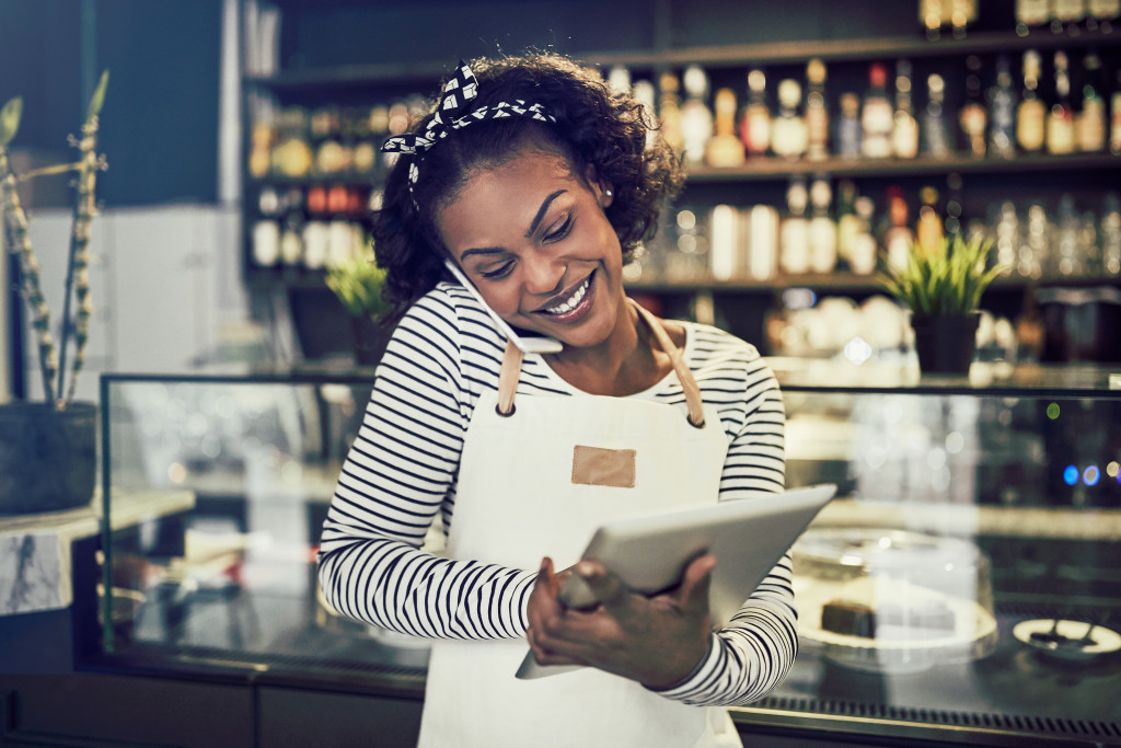 Young woman taking orders over the phone while using a tablet in front of the counter of her cafe.