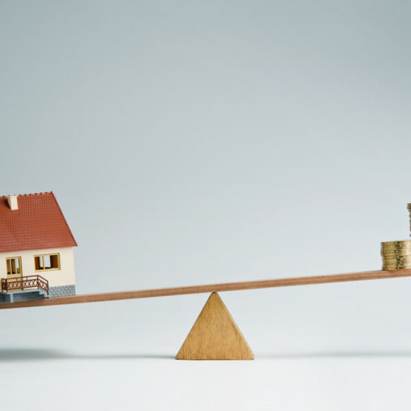 A concept photo of a house and a pile of coins on a seesaw with the house on the heavier side, symbolizing the increasing price of houses