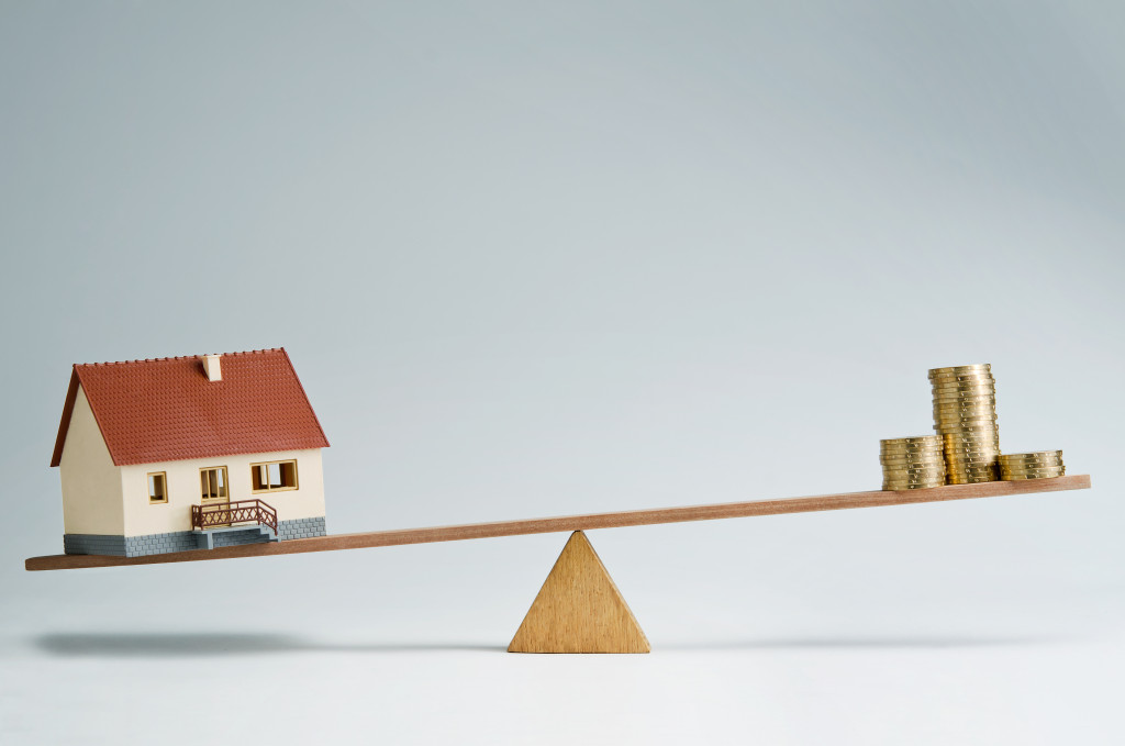 A concept photo of a house and a pile of coins on a seesaw with the house on the heavier side, symbolizing the increasing price of houses
